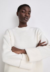 Knitted cream sweater with a round ribbed neckline and oversized, ribbed cuffs. Features a loose fit and textured fabric.