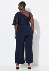 Mono navy con un diseño de un solo hombro, corsé de encaje y pantalones de pierna ancha con intrincados detalles de encaje a lo largo del costado.