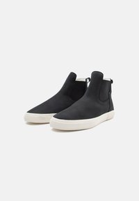 Botas negras hasta el tobillo hechas de material liso con suela de goma blanca, que presentan paneles laterales elásticos y tiradores para facilitar su uso.