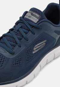 Skechers sportcipő, amely sötétkék lélegző hálós anyagból készült, szürke részletekkel és fehér gumitalppal, amely barázdákkal rendelkezik a tapadás érdekében.