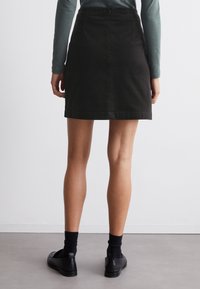 Zwarte denim mini-jurk met een aansluitende pasvorm, een ritssluiting aan de achterkant en een randdetail. Draag het met zwarte schoenen en sokken, tegen een neutrale achtergrond.