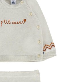 Jasno beżowy sweter z brązowym haftem "p'tit coeur", wyposażony w zapięcia na guziki i zygzakowaty wzór na rękawie. Miękka dzianinowa faktura.