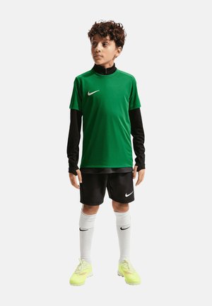 Junge mit lockigem Haar trägt ein grünes kurzärmeliges Trikot über einem schwarzen langärmeligen Oberteil, schwarze Shorts, weiße Socken und gelbe Sportschuhe.