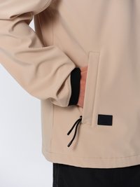 Beige lättviktig jacka med en mjuk textur, dragkedjeficka, justerbar dragsko och svarta detaljer vid ärmsluten.