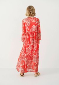 Lang rød maxi kjole med hvidt paisley print, lange ærmer og elastisk talje. Gennemsigtigt materiale og svajende kant. Sandaler bæres.