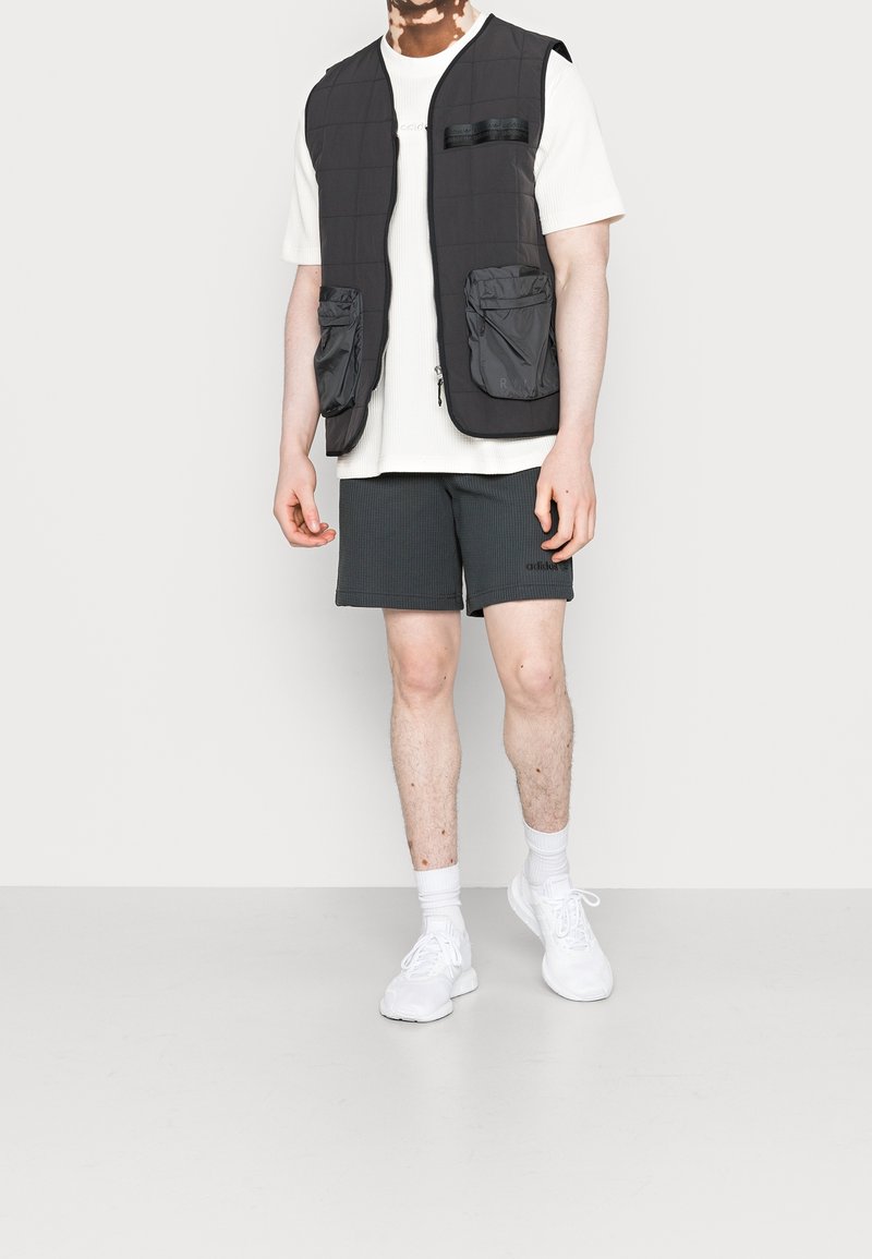 Černá zateplená vesta s dvěma předními kapsami, nošená přes bílou texturovanou košili a tmavé šortky. Model má na sobě bílé tenisky a ponožky.