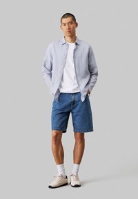 Camicia button-up a righe blu chiaro, T-shirt bianca, pantaloni corti in denim e sneakers beige. L'outfit presenta una vestibilità comoda e un design casual.