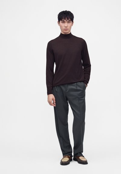 Homme portant un pull à col roulé marron foncé, un pantalon gris foncé et des mocassins marron et beige, debout avec une main dans la poche.