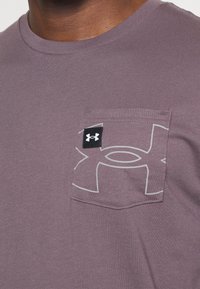 Camiseta de cuello redondo morada con un pequeño bolsillo en el pecho que presenta un gran logo débil de Under Armour y una etiqueta negra con el símbolo blanco de la marca.