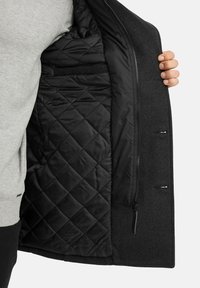 Schwarze Außenjacke mit einer gesteppten schwarzen Innenausstattung, ausgestattet mit einem Reißverschluss, einer Innentasche und einem eleganten Design. Sichtbares, strukturiertes Wollmaterial.