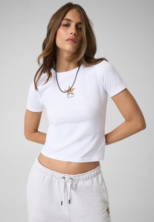 Femme aux longs cheveux bruns portant un t-shirt court blanc avec un petit motif étoile et un pantalon de survêtement gris clair, mains dans le dos.