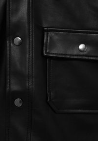 Gros plan sur une veste en cuir noire montrant une poche à rabat boutonnée et des boutons-pression argentés sur la patte de boutonnage avant.