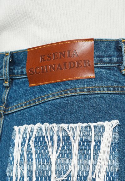 Denim jeans with a brown leather label featuring embossed text. Distressed detail with woven accents in lighter denim and white thread.