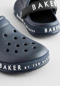 Mørkeblå crocs med ventilationshuller og en sort justerbar rem. Har hvid tekst "BAKER by TED BAKER" på siden. Gummimateriale.
