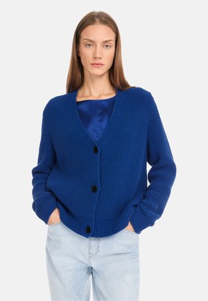 Blue knitted cardigan with a V-neck, long sleeves, and black buttons. Features a textured pattern and a relaxed fit. Paired with light jeans.