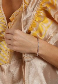 Silverarmband med klara ädelstenar, fäst på ett delikat band, som bärs på en hand bredvid ett mjukt, satinliknande, gyllene gult tyg.