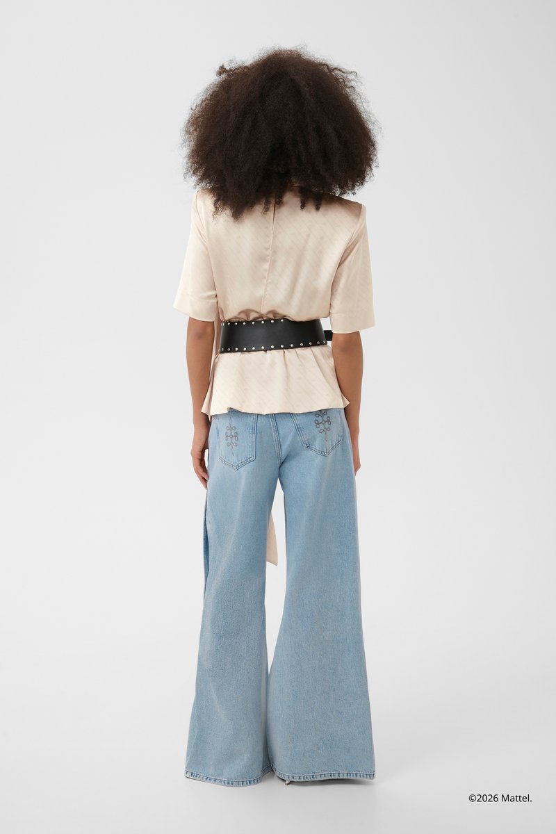 Kvinde med krøllet hår iført en beige kortærmet bluse, bredt sort bælte med nitter og lyseblå vidde jeans med syddelommer på bagsiden.