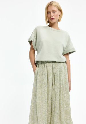 Blonde Frau trägt ein hellsalbegrünes kurzärmeliges Oberteil mit hochgekrempelten Ärmeln und einen hoch taillierten, gemusterten, fließenden Rock.