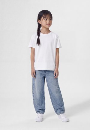Joven niña con el pelo trenzado, vistiendo una camiseta blanca, vaqueros sueltos azul claro y zapatillas blancas, de pie y mirando hacia un lado.