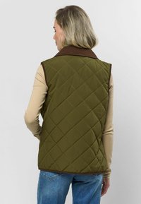Kurtka w kolorze oliwkowym, pikowana, z brązowym kołnierzem z kordurowej tkaniny, z wzorem w romby i wykończonym brzegiem, noszona na jasnobeżowej bluzce z długim rękawem.