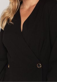 Svart omslagstyp plagg med djup V-ringning, långa ärmar och en guldfärgad accentknapp i midjan, tillverkat av ett slätt, texturerat tyg.