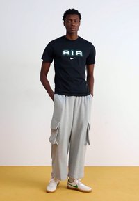 Juodas Nike marškinėlis su "AIR" logotipu, pilki laisvi sportiniai šortai su šoninėmis kišenėmis ir balti sportbačiai su žaliais akcentais. Paprastas fonas.