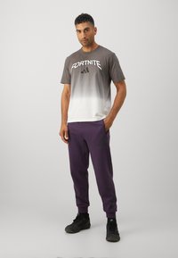 Tričko s prechodom farieb od sivej po bielu s nápisom "FORTNITE" a logom adidas, doplnené fialovými teplákmi s čiernymi akcentmi a vreckami. Čierne topánky.