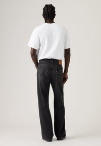 Μαύρο τζιν με ίσια γραμμή και καφέ δερμάτινο ετικέτα της μάρκας στο πίσω μέρος. Το μοντέλο φοράει απλό λευκό μπλουζάκι.