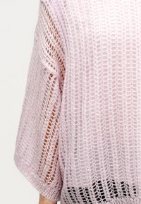 Ανοιχτό ροζ πουλόβερ με ανοιχτή πλέξη, μανίκια νυχτερίδας και χαλαρή ύφανση. Διαθέτει υφασμένο σχέδιο και φτάνει κάτω από τη μέση.