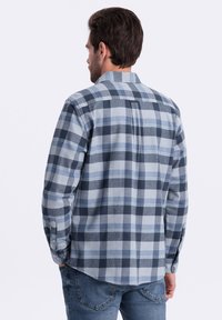 Camisa de franela a cuadros que presenta tonos de azul, gris y blanco. Mangas largas con puños abotonados y un pliegue en la parte posterior para mayor comodidad.