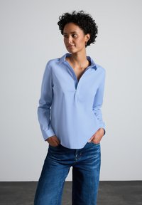 Camicia col celeste con colletto e una bottonatura, abbinata a jeans blu scuro. Tessuto morbido e vestibilità rilassata, senza fantasie o decorazioni.