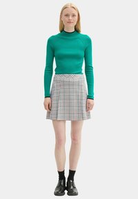 Grünes geripptes Langarmshirt kombiniert mit einem plissierten Kiltrock in karierten Blau-, Rot- und Cremetönen. Das Model trägt schwarze Socken und Schuhe.