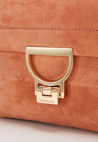 Beżowa torebka z zamszu z prostokątnym zapięciem w kolorze złotym, na którym wygrawerowano "COCCINELLE", z widocznym detalem przeszycia na górze.