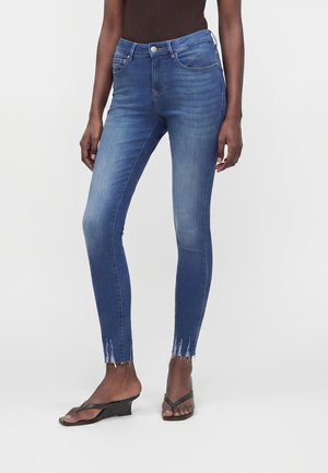 Woman wearing mid-rise blue skinny jeans with frayed hems and black wedge-strap sandals, standing against a plain background.
