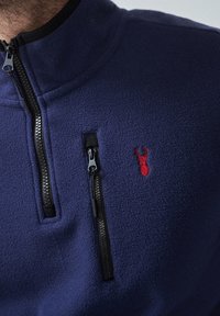 Marinefarbener Fleece-Pullover mit steigendem Kragen, ausgestattet mit einer schwarzen Reißverschlusstasche und einem roten gestickten Logo auf der Brust. Strukturierter Stoff.