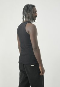 Schwarzes geripptes Tanktop, enganliegendes Design, Rückansicht, kombiniert mit dunklen Hosen, die ein Etikett an der Tasche haben. Neutraler Hintergrund.