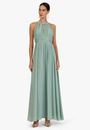 Frau, die ein ärmelloses, plissiertes, salbeigrünes Neckholder-Kleid trägt, dazu verzierte spitze Schuhe und hängende Ohrringe, steht vor einem weißen Hintergrund.