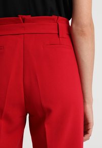 Close-up of a person wearing high-waisted red trousers with belt loops and back welt pockets, paired with a black top.