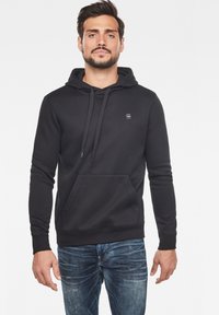 Svart bomullshoodie med en framficka, snörning på huvan och en liten logotyp på bröstet. Bärs med blå slitna jeans.