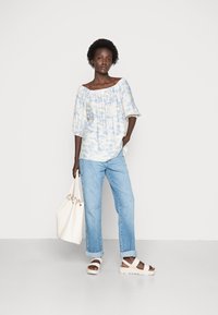 Top azul e branco com efeito tie-dye sem ombros e mangas bufantes, combinado com jeans claros de perna reta e sandálias brancas de plataforma, segurando uma bolsa tote branca.