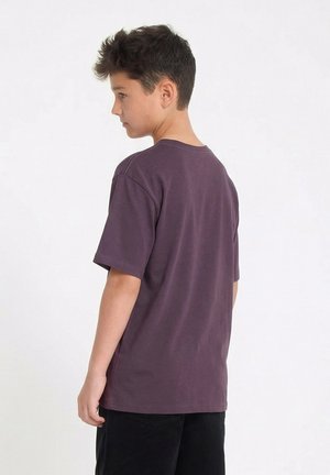 Un garçon aux cheveux courts et foncés se tient de profil, portant un t-shirt ample violet foncé et un pantalon noir, sur un fond clair uni.