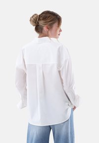 Weißes, langärmliges Hemd mit Knopfleiste, Rückenteil und Falten-Details, aus glattem Stoff gefertigt. Lange Ärmel mit geknöpften Manschetten. Getragen mit blauen Jeans.