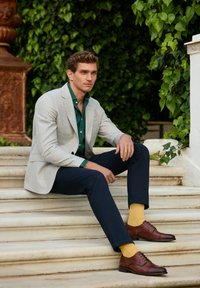 Blazer à carreaux gris clair sur une chemise verte, pantalon bleu marine et chaussures en cuir marron avec des chaussettes jaunes. Assis sur des marches avec un arrière-plan verdoyant.