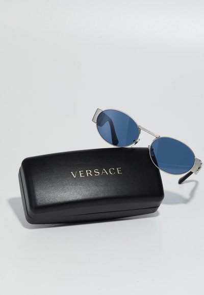Hõbedase metallraamiga prillid siniste läätsedega, puhates mustal ristkülikukujulisel karbil, millel on kuldne "VERSACE" logo.