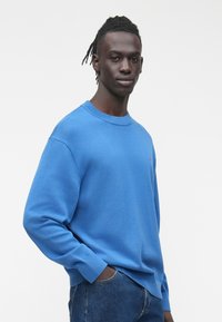 Mlad moški temnejše polti s kratkimi dreadloksi, oblečen v modro majico z okroglim ovratnikom in modre kavbojke, stoji pred belim ozadjem.