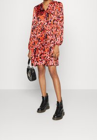Floral-patterned dress in shades of red and pink, features a V-neck, long sleeves, gathered waist, paired with black lace-up boots and a handbag.