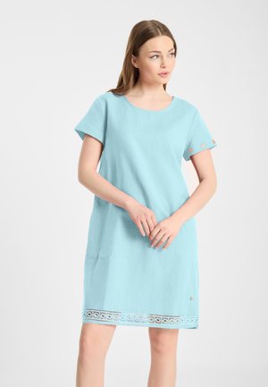 Jonge vrouw met lichtbruin haar die een lichtblauwe jurk met korte mouwen en een kanten rand aan de zoom draagt, kijkt naar de zijkant.