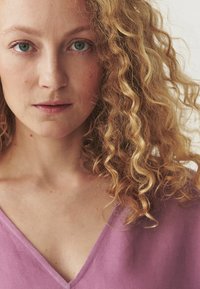 Portræt af en person med bølget, krøllet blond hår, iført en lavendel v-hals bluse. Stoffet ser blødt ud med en naturlig tekstur.