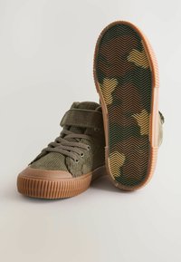 Baskets montantes en velours côtelé vert avec une semelle en caoutchouc beige, dotées de lacets et d'une bande Velcro. La semelle présente un motif camouflage avec un profil en chevrons.