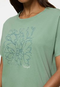 Zielona bawełniana koszulka z krótkim rękawem, z niebieskim kwiatowym wzorem w konturze i luźnym krojem. Zawiera detal z logo "Triumph" przy dolnym obszyciu.